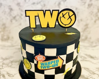 May include: A round cake with black and white checkered sides, topped with a yellow sign reading TWO and a smiley face icon. Decorations include yellow lightning bolts, additional smiley face icons, and a sign reading VANS WARPED TWO-UR.