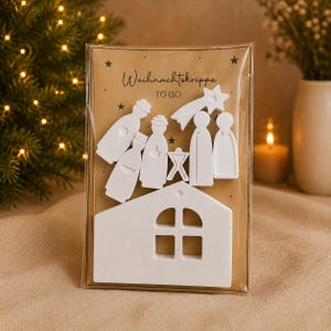 Könnte beinhalten: Ein Set aus weißen, flachen Holzfiguren für eine Krippe und einem hausförmigen Element, verpackt in einer transparenten Kunststoffhülle auf einer braunen Karte. Die Karte trägt den Text "Weihnachtskrippe TO GO" und kleine schwarze Sterne.