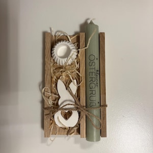 Könnte beinhalten: Osterdekoration aus Holz mit einem weißen Hasen-Ausschnitt, einer kleinen weißen Schale und einer grünen Kerze. Auf der Kerze steht der Text "Kleiner OSTERGRUB". Die Dekoration ist mit Bindfaden gebunden und mit Stroh gefüllt.