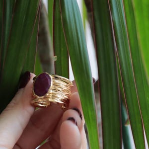 Puede incluir: Un anillo dorado con una gran piedra preciosa ovalada de color rojo oscuro. El anillo tiene un diseño texturizado y en capas. El fondo presenta hojas verdes y bordes rojos. El anillo es sostenido por una mano con esmalte de uñas oscuro.