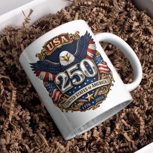 Puede incluir: Taza de cer&aacute;mica blanca con un dise&ntilde;o patri&oacute;tico para el 250 aniversario de los Estados Unidos de Am&eacute;rica. El dise&ntilde;o incluye un &aacute;guila, banderas americanas y el texto "USA 250 United States of America 1776-2026". La taza est&aacute; en una caja de regalo.