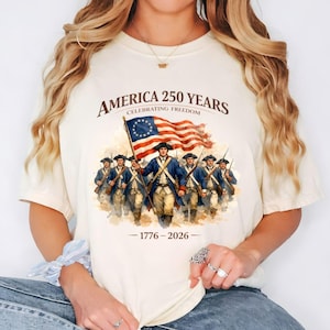 May include: A cream-coloured t-shirt with a graphic celebrating America's 250th anniversary. The design features the text "America 250 Years Celebrating Freedom" above an illustration of soldiers carrying the American flag, with the years 1776-2026 below.