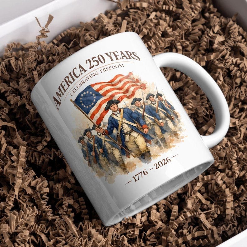 May include: White ceramic mug with a patriotic design. The mug features the text "AMERICA 250 YEARS CELEBRATING FREEDOM" and an illustration of soldiers carrying a flag. The date range "1776-2026" is also included.