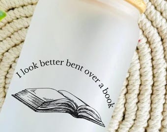 Może przedstawiać: Szklany kubek z matowego szkła z bambusową pokrywką. Kubek posiada czarną ilustrację otwartej książki oraz napis "I look better bent over a book". Złota naklejka w kształcie gwiazdy głosi "Hand pressed at home by me for you!"
