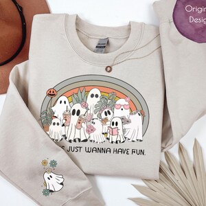 Könnte beinhalten: Beiges Sweatshirt mit einem Cartoon-Geister-Design. Das Design zeigt Geister mit Sonnenbrillen, Getränken und Büchern, vor einem Regenbogen. Der Text "Ghosts just wanna have fun" steht darunter. Ein Geist und Blumen sind auf dem Ärmel.