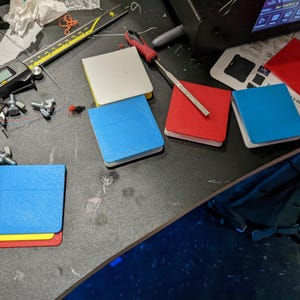Può includere: Un assortimento di oggetti quadrati colorati in rosso, blu, giallo e bianco è disposto su una superficie scura. Vari strumenti e hardware, tra cui un calibro e viti, sono sparsi intorno ai quadrati. La scena suggerisce un'officina o un ambiente artigianale.
