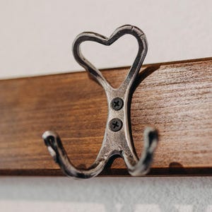 Könnte beinhalten: Ein metallener Kleiderhaken mit herzförmigem Oberteil und zwei unteren Haken, montiert auf einer Holzplatte. Der Haken ist mit zwei Schrauben befestigt. Das Holz hat einen warmen, braunen Ton, und die Wand ist weiß.