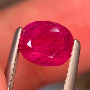 以下が含まれることがあります： 鮮やかなオーバルカットの宝石で、深く飽和したマゼンタピンクの色合いをしています。石の表面はファセット加工されており、光を反射して内部の模様やハイライトを作り出しています。銀色の金属製ピンセットでしっかりと保持されています。