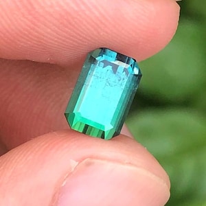 May include: A rectangular, emerald-cut gemstone in a vibrant teal hue, with a faceted surface. The gem is held between fingers, highlighting its clarity and brilliance. The facets reflect light, creating a sparkling effect.