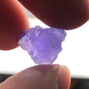 May include: A rough-cut, translucent purple gemstone is held between two fingers. The stone has an irregular shape with a crystalline structure, and the color varies from light to deep violet. The background is a bright, out-of-focus area.