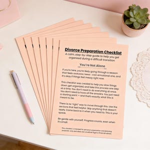 Op de afbeelding: Een stapel perzik-gekleurde "Divorce Preparation Checklist" papieren met tekst. Een pen met een lichtpaarse en roze schacht ligt op het witte oppervlak. Een kleine vetplant in een pot staat op de achtergrond.