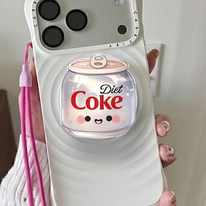 May include: White phone case with a Diet Coke design. The case features a cartoon Diet Coke can with a smiling face. A pink lanyard is attached to the case.