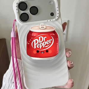 May include: A white phone case featuring a red Dr. Pepper can design with a smiling face. The case has a pink lanyard attached. The Dr. Pepper can has the brand name in white script.