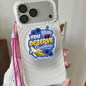 May include: White phone case with a graphic that says "You Deserve Sunshine" with a banana and a drink illustration. The case has a pink lanyard attached.