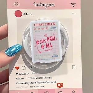 以下が含まれることがあります： 「JESUS PAID it ALL」のテキストが入った「Guest Check」デザインのピンク色のInstagramをテーマにしたスマホグリップ。ソーシャルメディアのアイコンと「520.1314 Likes」などのテキストが配置されたピンク色の背景に設定されています。