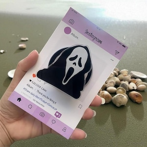 Può includere: Un pop socket di Ghostface in bianco e nero è mostrato su un finto post di Instagram. Il post è lilla chiaro e bianco, con il logo di Instagram e gli elementi dell'interfaccia. Il testo recita "Album. Grazie per aver messo mi piace" e include hashtag.
