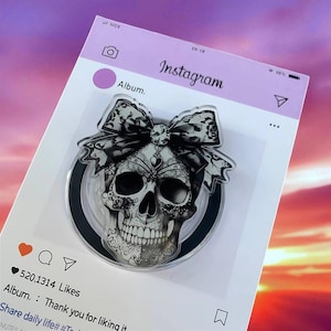 May include: A decorative skull-shaped accessory with a black and white bow. The skull has intricate floral designs and is set against a black circle. The background is a mock Instagram post with the text "Instagram" and "Album."