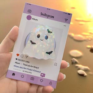 May include: A white ghost-shaped phone grip with a smiling face and three black bat accents. The grip is set against a light purple Instagram-themed background with the text "Album." and "Instagram."