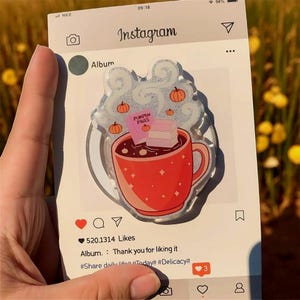 Może przedstawiać: Przezroczysta naklejka w stylu Instagrama z czerwoną filiżanką z gorącym napojem, dyniami i napisem "Pumpkin Pages". Naklejka jest trzymana w dłoni i ma interfejs mediów społecznościowych.