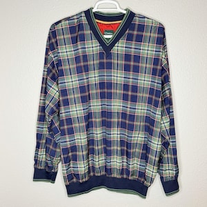 Könnte beinhalten: Ein langärmeliges kariertes Pulloverhemd in Marineblau, Grün und Rot. Das Hemd hat einen V-Ausschnitt mit marineblauer Einfassung und einen grün-marinefarbenen gestreiften Saum und Bündchen. Das Hemd hängt an einem weißen Kleiderbügel.