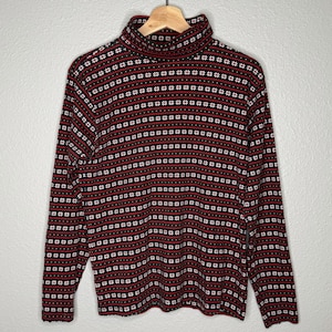 Pode incluir: Uma camisa de gola alta de manga comprida com um padrão repetitivo de listras horizontais vermelhas, pretas, brancas e verdes. A camisa está pendurada em um cabide de madeira contra um fundo neutro. O padrão inclui pequenas formas brancas em forma de flor.