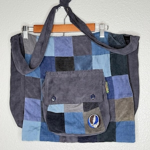 May include: A patchwork corduroy tote bag in shades of blue, gray, and black. The bag features a large front pocket with two button closures and a patch with a lightning bolt design. The bag hangs from a wooden hanger.