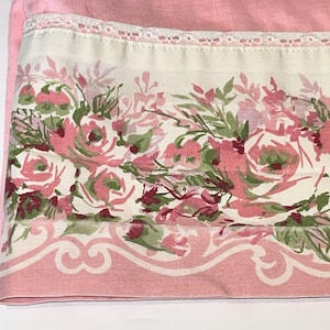 Puede incluir: Primer plano de una tela floral rosa y blanca. La tela presenta un borde rosa con un diseño floral blanco. El diseño floral incluye rosas rosas y hojas verdes. El borde superior tiene un ribete de encaje blanco.