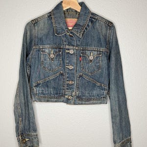May include: A cropped denim jacket in a medium wash blue. The jacket features a collared neckline, button-up closure, and multiple pockets. Silver-tone buttons and rivets add detail. A small red tag is visible on the chest pocket. The jacket is hanging on a wooden hanger.