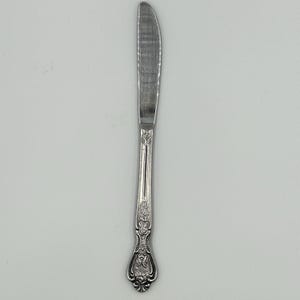 May include: A stainless steel butter knife with a curved blade and ornate floral detailing on the handle. The knife is designed for spreading butter or other soft spreads. The handle features a decorative, embossed pattern.