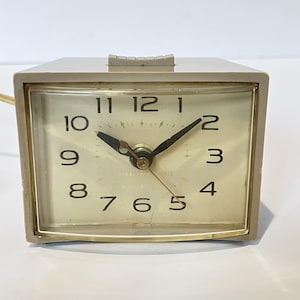 Puede incluir: Un reloj despertador eléctrico General Electric beige vintage. El reloj tiene una esfera rectangular con números y manecillas negras. La esfera del reloj está protegida por una cubierta de plástico transparente. El reloj tiene una caja de plástico beige y una palanca de ajuste de alarma montada en la parte superior.