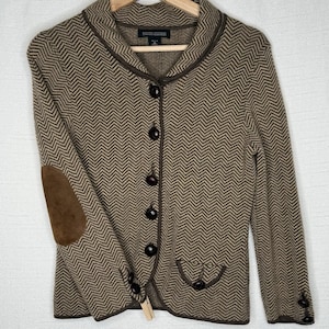 Puede incluir: Chaqueta marrón con estampado de espiga, cuello con solapas, cierre de botones y coderas de ante marrón. La chaqueta tiene un corte entallado y está colgada en una percha de madera.