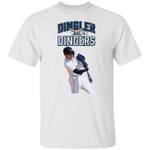 May include: A white short-sleeved t-shirt featuring a graphic of a baseball player swinging a bat. Above the player, the text reads DINGLER hits DINGERS in dark blue letters, accompanied by an illustration of two crossed bats and a baseball.