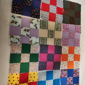 May include: A patchwork quilt top in progress, featuring a grid of colorful squares. The squares are made from various patterned fabrics, including floral prints, hummingbirds, and solid colors like red, blue, and green. The quilt is laid out on a white surface.