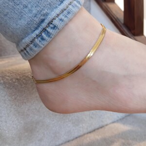 May include: A gold-colored herringbone chain anklet. The anklet is worn on the ankle, and the chain is flat and flexible. The anklet is a simple, elegant piece of jewelry.