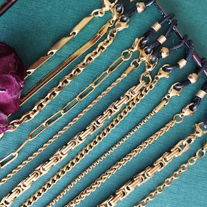 Peut inclure: Plusieurs chaînes de lunettes dorées de différents modèles, dont des chaînes à maillons, torsadées et à boîte. Les chaînes sont présentées sur une surface turquoise, avec une fleur violette séchée dans le coin. Les chaînes ont des boucles en caoutchouc noir.