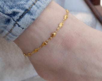 May include: A gold-colored anklet with a delicate chain design. The anklet features small, teardrop-shaped charms. The jewelry is worn on an ankle, with a denim-clad leg visible in the background.