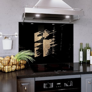 May include: Black kitchen backsplash with gold abstract design. The backsplash is installed behind a black stovetop and beneath a stainless steel range hood. A wire basket of potatoes and a small white bucket are visible.