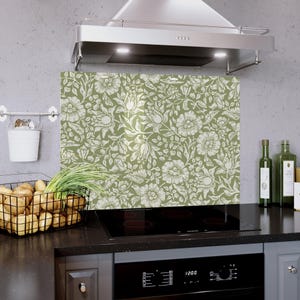 Pode incluir: Um painel de cozinha com padrão floral verde e branco. O design apresenta grandes flores e folhas estilizadas. O painel é instalado atrás de um fogão preto e em frente a um exaustor de aço inoxidável.