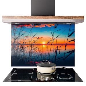 Op de afbeelding: Een keukenafzuigkap met houten rand en glazen paneel met een zonsondergang boven een meer met riet. Een zwarte kookplaat met een crèmekleurige pan met deksel en zilveren handvat.