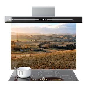 May include: A black and silver range hood, branded "PRISMA", is positioned above a black glass hob with circular heating zones. A white saucepan rests on the hob. The background features a scenic landscape.