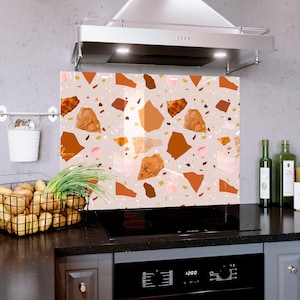 May include: A decorative kitchen backsplash with a terrazzo pattern. The design features a light beige background with various shapes in shades of brown, orange, and pink. The backsplash is installed above a black stovetop and below a stainless steel range hood.