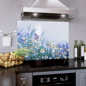 Può includere: Pannello paraschizzi da cucina in vetro raffigurante un campo di margherite bianche, fiori selvatici viola e una farfalla. L'immagine ha uno sfondo blu tenue. Il pannello è installato sopra un piano cottura nero e sotto una cappa aspirante in acciaio inossidabile.