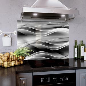 May include: A black and white abstract glass kitchen backsplash with a wavy design. The backsplash is installed behind a black stovetop and beneath a stainless steel range hood. A wire basket of potatoes and a small white bucket hang on the left.