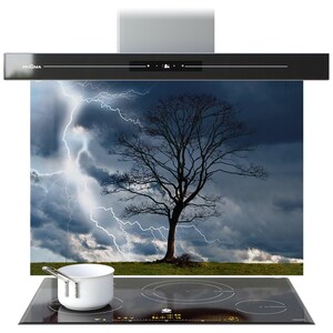 May include: A contemporary kitchen extractor fan in black and silver. It has a digital display and is positioned above a glass panel depicting a lightning storm and a bare tree. A black hob with a white saucepan is in the foreground.