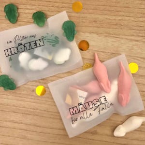 以下が含まれることがあります： 2つの小さなセロハン袋。1つは緑色のカエル型キャンディーとテキスト「ein Polster aus KR&Ouml;TEN」、もう1つはピンク色のネズミ型キャンディーとテキスト「M&Auml;USE f&uuml;r alle F&auml;lle」が入っています。散らばった紙吹雪。