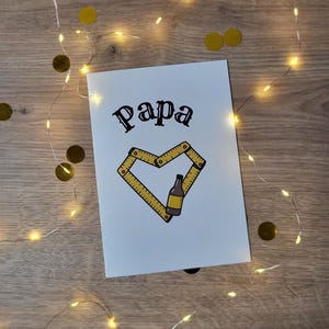 May include: A white greeting card with the word "Papa" in black script. A yellow ruler forms a heart shape around a beer bottle illustration. The card is decorated with gold confetti and fairy lights.