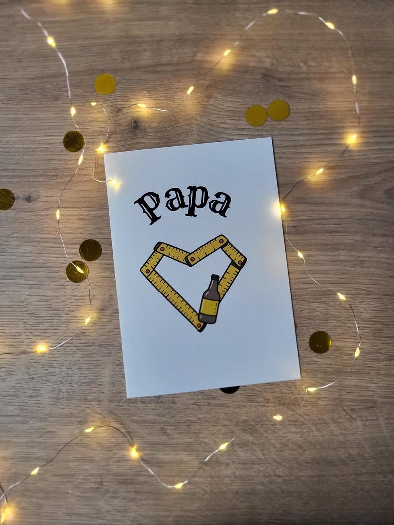 May include: A white greeting card with the word "Papa" in black script. A yellow ruler forms a heart shape around a beer bottle illustration. The card is decorated with gold confetti and fairy lights.