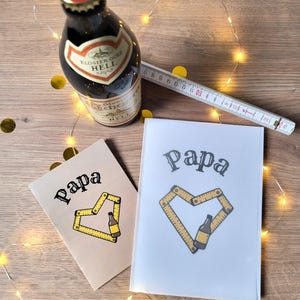 May include: Two greeting cards with the word "Papa" and a heart-shaped design made of a measuring tape and a beer bottle. A bottle of beer and a measuring tape are also present.