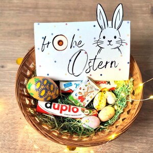 May include: A woven basket filled with Easter treats, including a decorated egg, chocolate bars, and small candies. A sign with a bunny illustration and the words "Frohe Ostern" is in the basket. The basket is on a wooden surface.