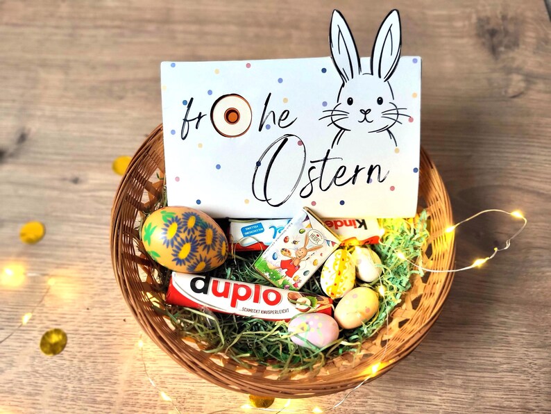 May include: A woven basket filled with Easter treats, including a decorated egg, chocolate bars, and small candies. A sign with a bunny illustration and the words "Frohe Ostern" is in the basket. The basket is on a wooden surface.